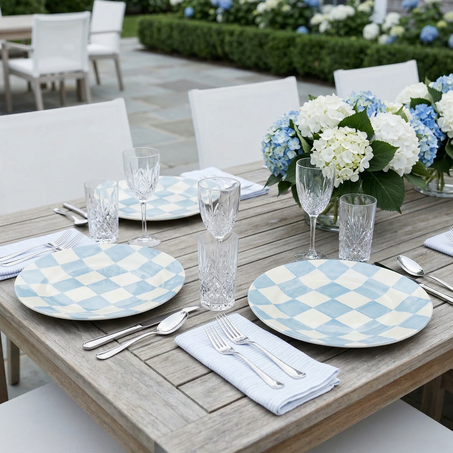 Light blue checkered melamine dinner plates on an outdoor wooden table with glassware and white hydrangea centerpiece.